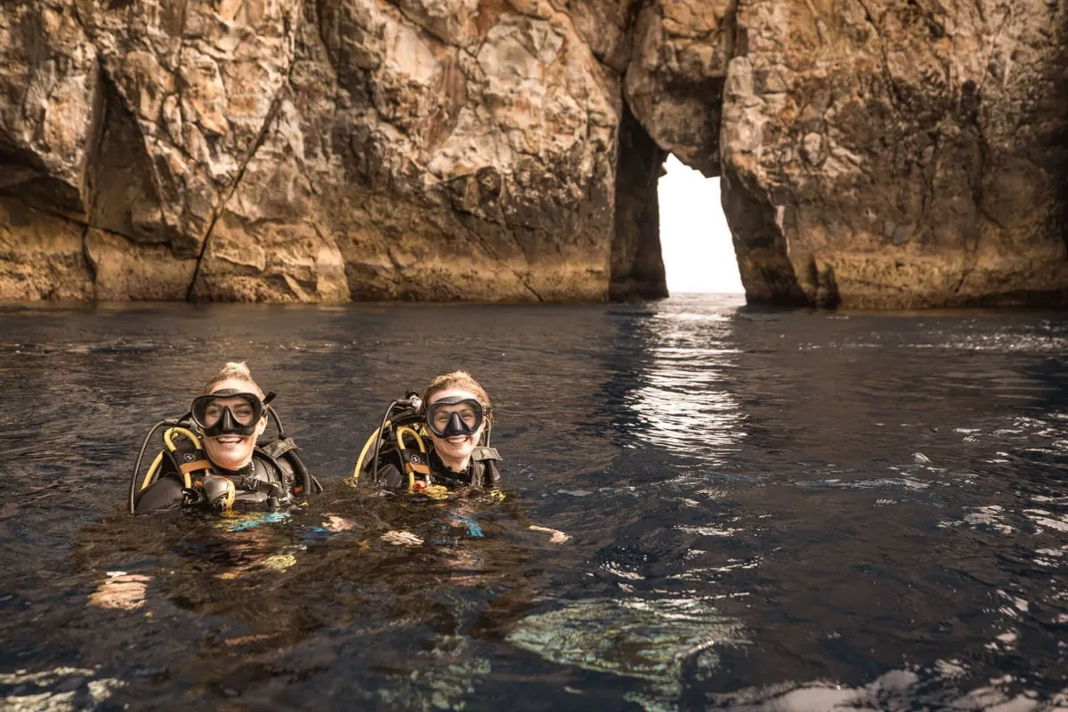 Two scuba divers floating in the water in front of a rocky sea arch at Poor Knights Islands, New Zealand.